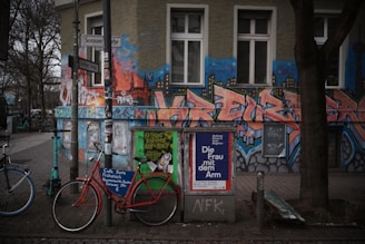 An urban street scene featuring a vibrant, graffiti-covered building exterior with bold, stylized letters and cityscape motifs. In front, bicycles are parked beside a pole laden with posters, including an advertisement for a cultural event. Trees with bare branches line the sidewalk, and the surroundings convey a mix of art and everyday urban life.