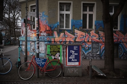 An urban street scene featuring a vibrant, graffiti-covered building exterior with bold, stylized letters and cityscape motifs. In front, bicycles are parked beside a pole laden with posters, including an advertisement for a cultural event. Trees with bare branches line the sidewalk, and the surroundings convey a mix of art and everyday urban life.