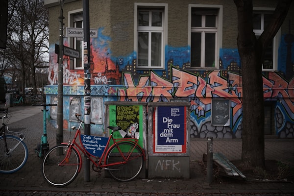 An urban street scene featuring a vibrant, graffiti-covered building exterior with bold, stylized letters and cityscape motifs. In front, bicycles are parked beside a pole laden with posters, including an advertisement for a cultural event. Trees with bare branches line the sidewalk, and the surroundings convey a mix of art and everyday urban life.
