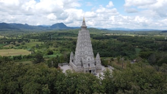 A large, intricately designed temple structure is set amidst lush greenery and rolling hills. The temple features detailed carvings and a prominent central spire topped with a golden pinnacle, standing out in the natural landscape. The sky is partly cloudy, adding a serene backdrop to the scene.