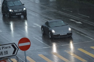 Two cars are driving on a wet, reflective road on a rainy day. A black SUV and a dark-colored sports car are visible, along with a no-entry traffic sign and a directional sign with text. The road has visible lane markings and a pedestrian crossing.