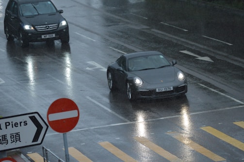 Two cars are driving on a wet, reflective road on a rainy day. A black SUV and a dark-colored sports car are visible, along with a no-entry traffic sign and a directional sign with text. The road has visible lane markings and a pedestrian crossing.