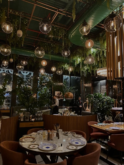 A stylish restaurant interior with a lush, green decor theme. Hanging pendant lights and ceiling foliage create a cozy, intimate atmosphere. The tables are elegantly set with patterned plates and glassware. People are seen working or possibly attending to guests, and the seating areas feature plush chairs.