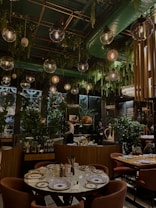 A stylish restaurant interior with a lush, green decor theme. Hanging pendant lights and ceiling foliage create a cozy, intimate atmosphere. The tables are elegantly set with patterned plates and glassware. People are seen working or possibly attending to guests, and the seating areas feature plush chairs.
