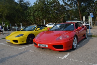 Two sports cars are parked side by side in a parking lot. There is a red car with license plate XF 2994 and a yellow car with license plate RP 2527. Surrounding the cars are trees and other parked vehicles. The day appears sunny, casting clear shadows on the ground.