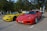 Two sports cars are parked side by side in a parking lot. There is a red car with license plate XF 2994 and a yellow car with license plate RP 2527. Surrounding the cars are trees and other parked vehicles. The day appears sunny, casting clear shadows on the ground.