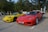 Two sports cars are parked side by side in a parking lot. There is a red car with license plate XF 2994 and a yellow car with license plate RP 2527. Surrounding the cars are trees and other parked vehicles. The day appears sunny, casting clear shadows on the ground.