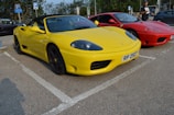 Two sports cars are parked side by side in a parking lot. The car in the foreground is bright yellow with a convertible top, while the car behind it is red. The yellow car has a visible license plate with the number 'RP 2527'. A person stands near the red car, holding something in their hand.