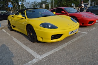 Two sports cars are parked side by side in a parking lot. The car in the foreground is bright yellow with a convertible top, while the car behind it is red. The yellow car has a visible license plate with the number 'RP 2527'. A person stands near the red car, holding something in their hand.