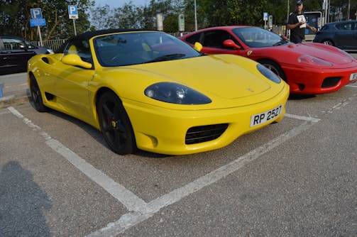 Two sports cars are parked side by side in a parking lot. The car in the foreground is bright yellow with a convertible top, while the car behind it is red. The yellow car has a visible license plate with the number 'RP 2527'. A person stands near the red car, holding something in their hand.