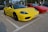 Two sports cars are parked side by side in a parking lot. The car in the foreground is bright yellow with a convertible top, while the car behind it is red. The yellow car has a visible license plate with the number 'RP 2527'. A person stands near the red car, holding something in their hand.