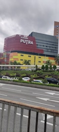 A multi-story building with a modern design, featuring a vibrant facade in red, yellow, and black. The structure is prominently labeled 'Sunway Putra' and is located near a busy road. Several cars can be seen on the road and a landscaped area with small trees and shrubs is in the foreground. The sky appears overcast, hinting at cloudy weather.