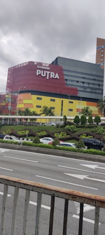 A multi-story building with a modern design, featuring a vibrant facade in red, yellow, and black. The structure is prominently labeled 'Sunway Putra' and is located near a busy road. Several cars can be seen on the road and a landscaped area with small trees and shrubs is in the foreground. The sky appears overcast, hinting at cloudy weather.