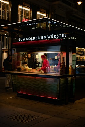 A nighttime scene featuring a street food stall with a bright neon sign reading 'Zum Goldenen W&uuml;rstel'. The stall is illuminated with warm lights, and a person wearing a red cap and jacket is preparing food. Another person stands outside the stall, seemingly waiting. The ambiance suggests a bustling city evening.