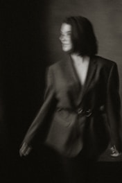 A blurred black-and-white photograph of a person wearing an oversized blazer. The individual has short hair and stands against a dark, partially lit background, creating a moody and indistinct atmosphere.