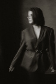 A blurred black-and-white photograph of a person wearing an oversized blazer. The individual has short hair and stands against a dark, partially lit background, creating a moody and indistinct atmosphere.