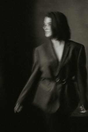 A blurred black-and-white photograph of a person wearing an oversized blazer. The individual has short hair and stands against a dark, partially lit background, creating a moody and indistinct atmosphere.