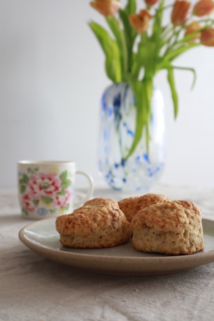 A ceramic plate holds three golden-brown scones, placed on a textured linen tablecloth. In the background, there is a colorful floral-patterned teacup alongside a glass vase containing fresh orange tulips with green leaves.