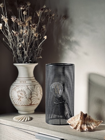 A decorative arrangement featuring a white ceramic vase with intricate floral patterns, holding dried flowers with earthy tones. Adjacent to the vase is a black perforated cylindrical lamp and two seashells, all placed on a wooden surface.