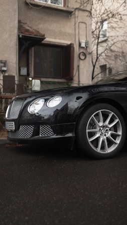 A luxury black car with a distinctive grille and shiny alloy wheels is parked close to a residential building. The building's exterior is textured, with visible windows and air conditioning units. The ground is wet, possibly from recent rain, and the surroundings include bare tree branches, suggesting an overcast day.