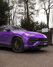 A vibrant purple luxury SUV is parked on a paved surface, surrounded by tall trees and a building with a wooden facade. The vehicle boasts a sleek design with large black alloy wheels and yellow brake calipers, enhancing its sporty appearance.