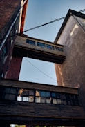 Two industrial buildings are connected by wooden skybridges with multiple windows. Overhead wires stretch across the scene, while the afternoon light casts shadows on the textured surfaces.