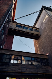 Two industrial buildings are connected by wooden skybridges with multiple windows. Overhead wires stretch across the scene, while the afternoon light casts shadows on the textured surfaces.