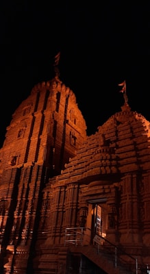 An intricately designed temple with detailed carvings adorning its facade, illuminated warmly against a dark night sky. The structure features tall towers with flags at the top, highlighting its architectural grandeur.