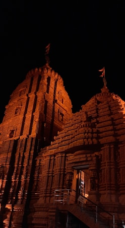 An intricately designed temple with detailed carvings adorning its facade, illuminated warmly against a dark night sky. The structure features tall towers with flags at the top, highlighting its architectural grandeur.