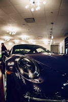 A sleek, dark-colored sports car with large circular headlights is prominently displayed indoors. The setting is well-lit with multiple modern ceiling lights casting reflections on the car's glossy surface. A person in the background adds to the sophisticated ambiance of the showroom.