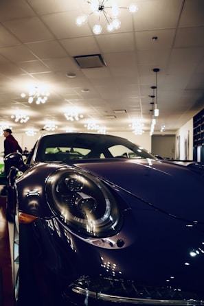 A sleek, dark-colored sports car with large circular headlights is prominently displayed indoors. The setting is well-lit with multiple modern ceiling lights casting reflections on the car's glossy surface. A person in the background adds to the sophisticated ambiance of the showroom.