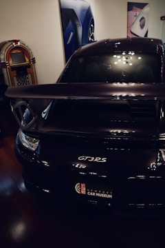 A sleek black car with a prominent rear spoiler is parked indoors. On the left, there is a vintage jukebox, and the walls are adorned with large automotive-themed photographs.