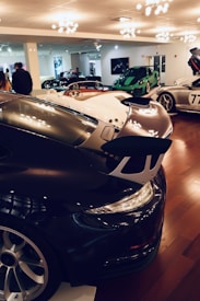 A car showroom displays several high-end sports cars, including sleek models in various vibrant colors. The space is well-lit with modern lighting fixtures, and there are a few people observing the vehicles.