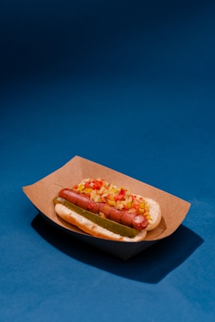 A hot dog in a bun topped with diced onions and pickles is placed in a brown paper tray. The background is a solid deep blue, creating a vibrant contrast.