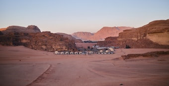 A desert landscape with a small settlement of white dome tents in the center, surrounded by towering rocky cliffs. The sky is clear, and the terrain is sandy with a few tracks leading to the settlement.