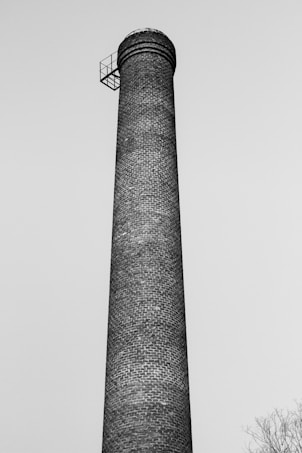 A tall brick chimney structure extending high into the sky. There is a small metal platform with a railing at the top of the chimney.