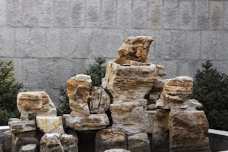 A composition of large, rugged stone blocks stacked together, surrounded by greenery such as pine trees. The textured surface of the stones exhibits layers of various earthy colors, and the background features a gray, tiled wall.