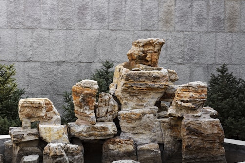 A composition of large, rugged stone blocks stacked together, surrounded by greenery such as pine trees. The textured surface of the stones exhibits layers of various earthy colors, and the background features a gray, tiled wall.