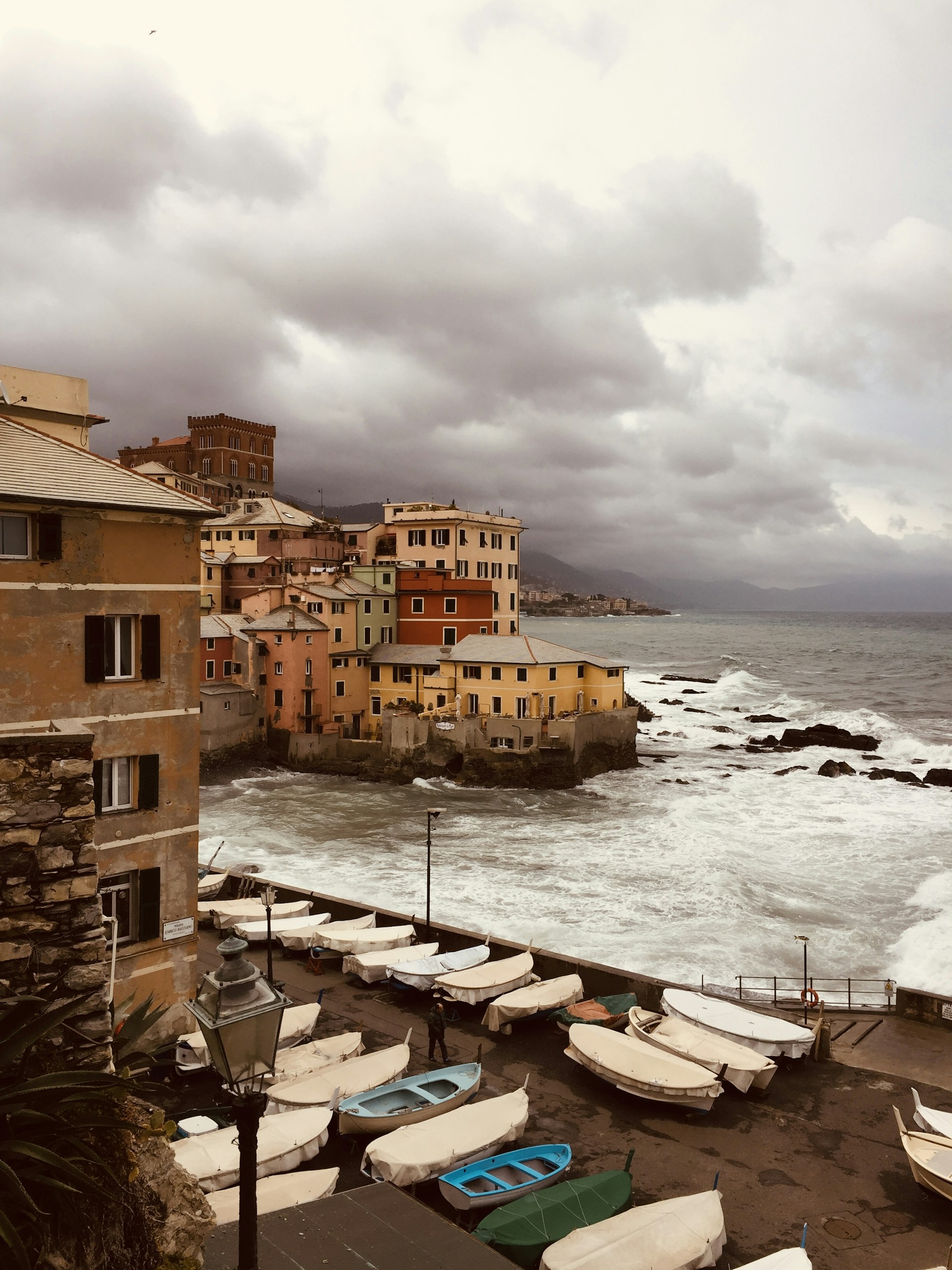 A coastal scene featuring a quaint seaside village with colorful buildings, a choppy sea with crashing waves, and a cloudy sky. Boats are lined up along the waterfront, adding to the scenic view.