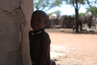 A young child stands next to a weathered, cracked mud wall. Several beaded necklaces adorn their neck, and the background features a sunlit outdoor scene with trees and traditional structures that resemble huts or fences.