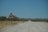 A dirt road stretches into the distance through a dry savannah landscape with low shrubs and distant hills. On the left side of the road, there is a triangular road sign depicting a leaping antelope, warning of potential wildlife crossings.