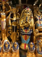 The image displays a collection of ornate Egyptian statues and artifacts featuring vibrant colors and intricate details. Prominent figures include pharaohs and deities adorned in gold, blue, and red.