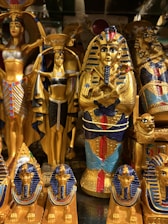 The image displays a collection of ornate Egyptian statues and artifacts featuring vibrant colors and intricate details. Prominent figures include pharaohs and deities adorned in gold, blue, and red.