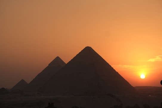 A desert landscape featuring three large pyramids against a vibrant orange sunset. The sun is low on the horizon, casting a warm glow over the scene. The sky gradually transitions from a deep orange near the sun to a darker shade further away.