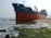 A large cargo ship with the identification 'HAIPHONG 45' is stranded on a sandy shoreline. The hull is mostly red with a blue stripe near the top. The water around the ship is relatively calm, with some waves crashing gently against the shore. On the beach, there is green seaweed or moss present on the sand and rocks. The sky is clear with a subtle haze.