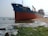 A large cargo ship with the identification 'HAIPHONG 45' is stranded on a sandy shoreline. The hull is mostly red with a blue stripe near the top. The water around the ship is relatively calm, with some waves crashing gently against the shore. On the beach, there is green seaweed or moss present on the sand and rocks. The sky is clear with a subtle haze.