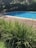 A peaceful outdoor scene featuring a swimming pool with clear blue water, surrounded by lush green vegetation and a wooden deck. A metal and fabric poolside chair is positioned next to the pool on the deck. Tall grass in the foreground adds a natural touch, while flowering bushes and trees in the background contribute to the serene atmosphere.