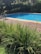 A peaceful outdoor scene featuring a swimming pool with clear blue water, surrounded by lush green vegetation and a wooden deck. A metal and fabric poolside chair is positioned next to the pool on the deck. Tall grass in the foreground adds a natural touch, while flowering bushes and trees in the background contribute to the serene atmosphere.
