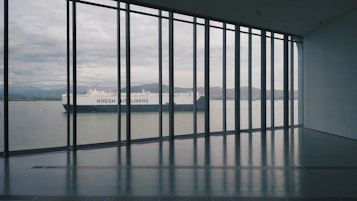 A large ship is visible through the floor-to-ceiling windows of a modern building. The ship, with the words 'H&ouml;egh Autoliners' on its side, is situated on a calm body of water with mountain ranges visible in the distance. The interior of the building features a dark, glossy floor and a minimalist design.