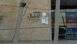 A wall with a large sign for The Children's Museum in Arabic and English, featuring a simple drawing of a child and an adult in traditional clothing. The background is made of brick-like material, and the entrance with glass panels is visible.